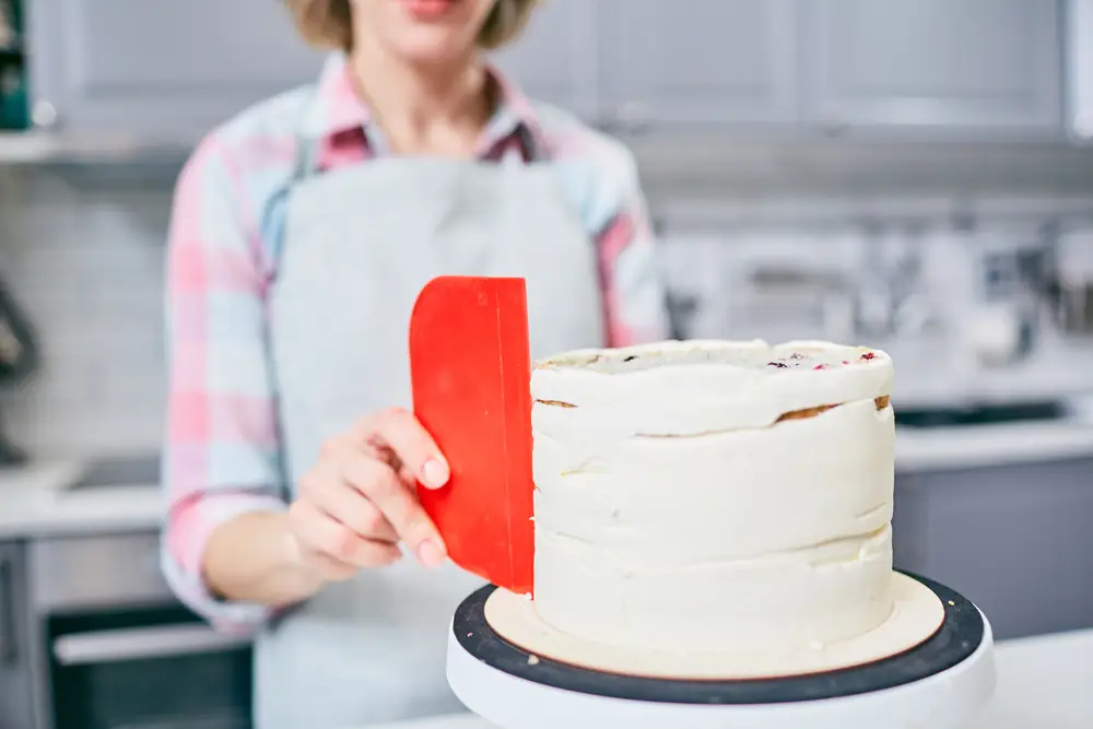 Confeiteira alisando a lateral de um bolo branco com espátula rosa sobre o prato giratório